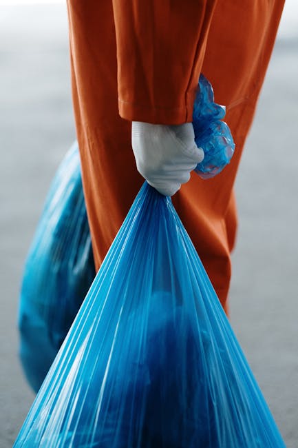 A person wearing orange protective overalls and a white glove is holding a large, semi-transparent blue plastic bag filled with waste materials. The bag appears to be tightly gathered and secured at the top, with some visible contents inside, possibly indicating waste or rubbish for disposal. The individual is standing outdoors on a concrete or asphalt surface, with the background showing a blurred, neutral-toned environment that suggests a waste disposal site or collection area. The scene emphasizes private waste handling or rubbish removal activities, typical of an independent disposal operation conducted by waste management services like Waste Disposal Fulham. The focus on the hand gripping the bag and the bag's size and material underscore the nature of rubbish collection, highlighting logistics involved in on-site clearance or alternative waste collection methods outside of standard municipal services.