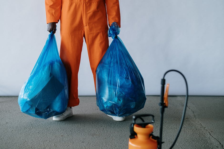 A person wearing orange protective overalls and a white glove is holding a large, semi-transparent blue plastic bag filled with waste materials. The bag appears to be tightly gathered and secured at the top, with some visible contents inside, possibly indicating waste or rubbish for disposal. The individual is standing outdoors on a concrete or asphalt surface, with the background showing a blurred, neutral-toned environment that suggests a waste disposal site or collection area. The scene emphasizes private waste handling or rubbish removal activities, typical of an independent disposal operation conducted by waste management services like Waste Disposal Fulham. The focus on the hand gripping the bag and the bag's size and material underscore the nature of rubbish collection, highlighting logistics involved in on-site clearance or alternative waste collection methods outside of standard municipal services.