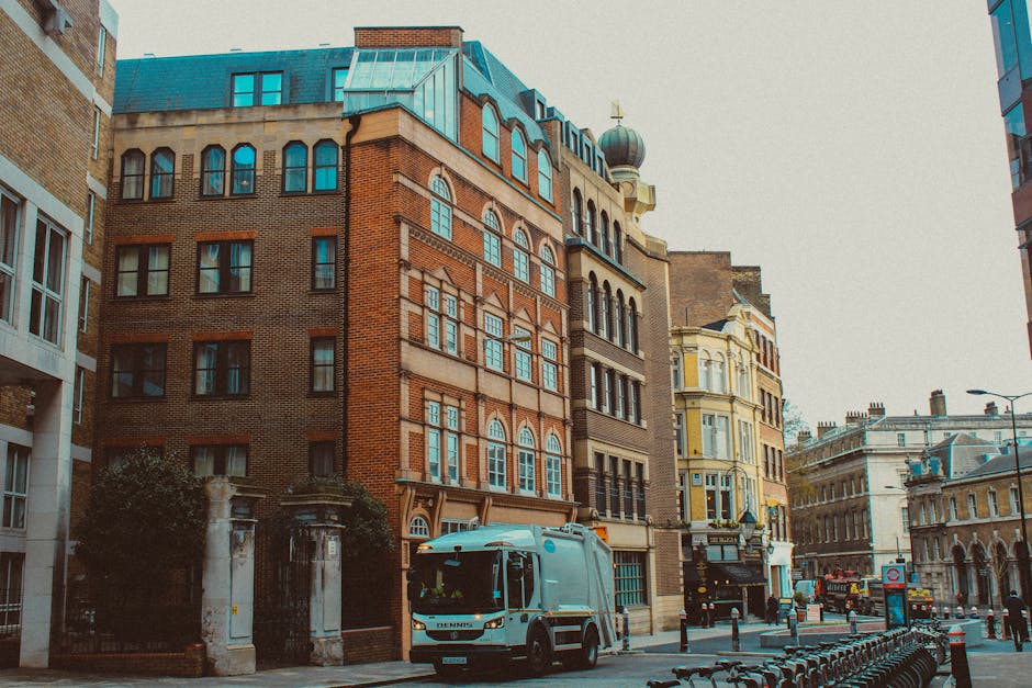 The image depicts a city street scene featuring a row of historic and modern buildings with varied architectural styles, including brick facades, arched windows, and decorative cornices, all under a clear sky. In the foreground, a grey rubbish collection truck from Waste Disposal Fulham is parked on the curb, positioned adjacent to a designated waste storage area. The truck has a modern, clean appearance with a compact design suitable for urban service access. Surrounding the truck, there are a few black wheelie bins and refuse containers aligned along the sidewalk, likely awaiting collection. The street itself is lined with parked bicycles, metal bollards, and a pedestrian pathway, with some greenery visible behind a wrought-iron gate in front of a building. The ambient lighting suggests daytime with natural light illuminating the scene, underscoring the urban context where private waste collection services may be used as part of alternative rubbish disposal arrangements in Fulham. Overall, the scene emphasizes the interaction of urban architecture and waste management logistics within a busy city environment, aligning with the themes of rubbish removal and on-site clearance services offered by Waste Disposal Fulham.
