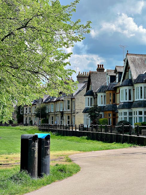 A row of Victorian-style terraced houses with bay windows and pitched roofs, situated along a residential street under a partly cloudy sky. In the foreground, two black outdoor rubbish and recycling bins are placed on a grassy verge next to a paved footpath. A green-leafed tree extends its branches over the bins, providing some shade. Behind the bins, a black railing runs parallel to the houses, separating the sidewalk from small front gardens. The houses feature chimney stacks and display a mix of brickwork and painted finishes, with some including decorative wooden trim around the windows. The scene suggests a peaceful suburban environment, typical of urban residential areas where local waste disposal or private rubbish collection services, such as those provided by Waste Disposal Fulham, are used for regular waste management in this community.