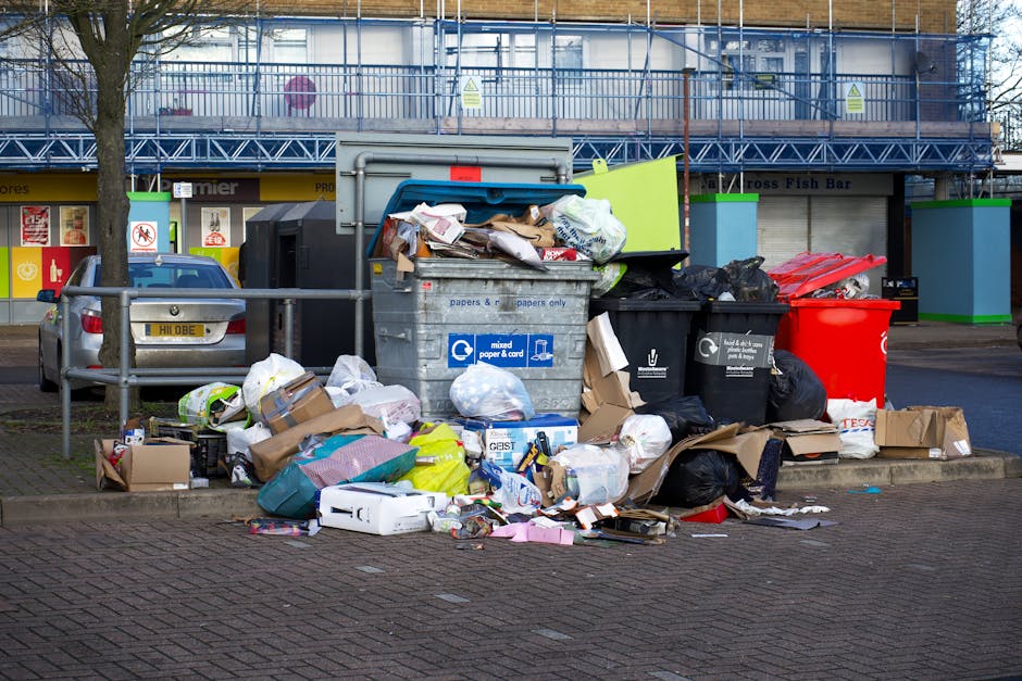 A large pile of mixed waste, including cardboard boxes, plastic bags, paper debris, and discarded packaging materials, is overflowing from a grey metal recycling bin and surrounding black and red waste containers, situated on a paved urban sidewalk near parked cars. The recycling bin, positioned centrally, is partially open with various papers and packaging spilling out onto the ground in front. Nearby, black rubbish bags are stacked and some are torn, with additional loose waste scattered across the area. The background features a commercial building with a blue temporary fencing and scaffolding, alongside shop signage indicating different retail outlets. A tree with a bare trunk is located to the left of the waste pile, and a metal railing runs parallel to the pavement, separating the waste area from a parking area where a silver vehicle is parked. The scene appears to be an instance of unmanaged or overflowing refuse, highlighting the importance of regular waste collection and the services offered by waste disposal companies such as Waste Disposal Fulham in maintaining urban cleanliness and proper rubbish handling.