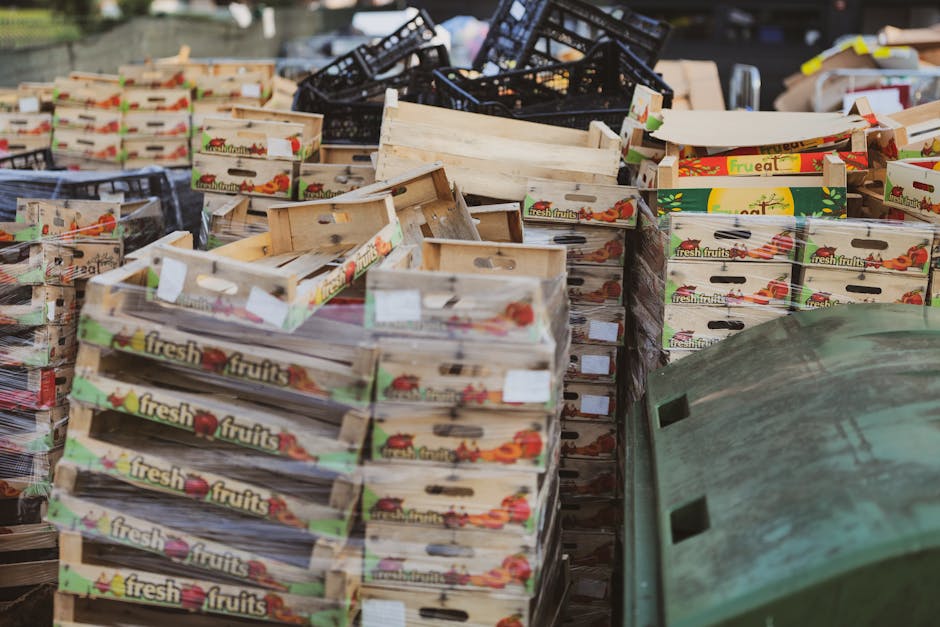A large outdoor pile of cardboard fruit boxes, primarily labeled 'fresh fruits' with colorful images of apples, tomatoes, and other produce, stacked and scattered across a paved area. Some boxes are damaged or partially collapsed, with a mix of open and closed containers. Behind the boxes, there are black plastic crates piled irregularly, and in the foreground, a weathered green plastic container with a slightly textured surface. The scene is illuminated by natural daylight, suggesting an outdoor waste or rubbish collection setting, with the environment indicating an area designated for private waste disposal or bulk rubbish removal, consistent with services provided by Waste Disposal Fulham for handling unwanted household or commercial waste materials.