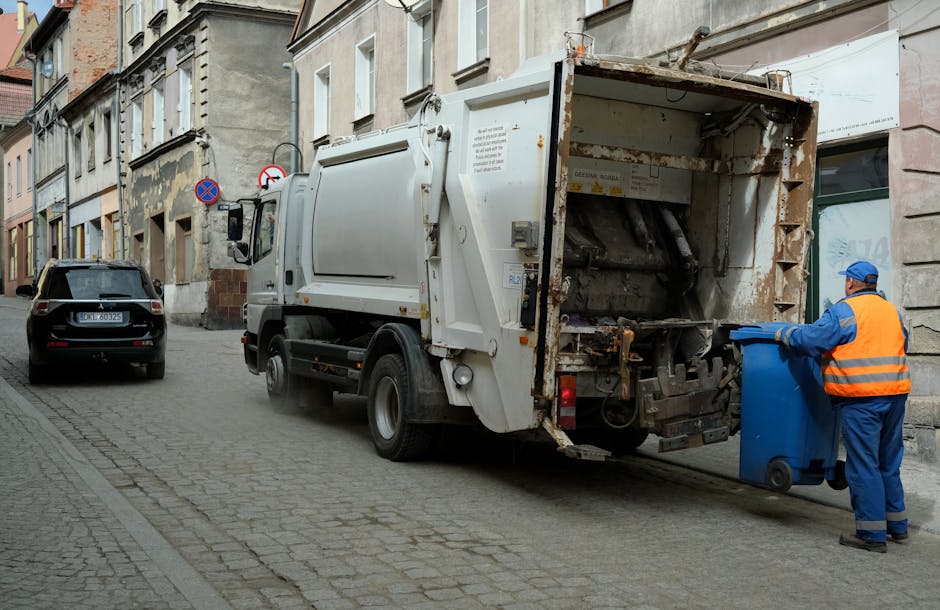 A waste collection truck with a white exterior and a rusted, open rear hopper is positioned on a narrow cobblestone street, actively engaged in rubbish removal. A worker dressed in blue overalls, a bright orange high-visibility vest, and a blue cap is seen beside the truck, emptying a blue wheeled bin into the hopper. The street is lined with multi-story residential buildings featuring aged, textured facades in neutral tones, some with visible signs of wear and weathering. A black car is parked further down the street, and traffic signs, including a no parking and no stopping sign, are affixed to the building walls. The truck's front cab and side mirror are intact, and the environment appears to be during daylight with diffused natural light. The scene exemplifies private waste collection services replacing or supplementing local authority rubbish collection, with emphasis on on-site rubbish removal from residential premises, compatible with services like those offered by Waste Disposal Fulham.