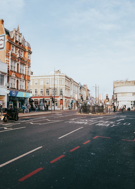 A daytime street scene in an urban area with a row of multi-story buildings featuring Victorian and modern architectural styles. The buildings have detailed facades, with some brickwork and decorative elements painted in light and dark tones. The street has marked lanes, including a designated lane for buses and a bus stop with sheltered seating and a timetable display. Several pedestrians are walking along the sidewalks, and a motorcycle is parked beside the kerb. In the background, there are traffic lights, street signs, and utility poles with overhead wires. The sky is partly cloudy with a soft, natural light illuminating the scene. The image also shows a section of an empty road with red dashed lines indicating a bus lane or special restriction zone, reflecting the type of private or independent waste collection operations that sometimes occur in urban environments, as seen by the proximity of Waste Disposal Fulham’s services in similar settings.