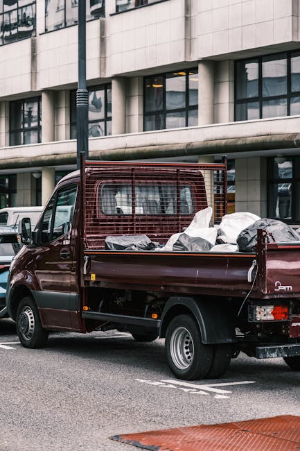 A dark red flatbed utility vehicle parked on a city street in front of a modern, multi-story building with large, tinted windows and a light-colored facade. The truck's cargo area is loaded with various mixed waste materials, including black and white plastic bags, some of which appear to contain household or construction rubbish. The bags and loose debris are piled unevenly in the truck bed, with some items leaning against the metal rear wall, which features a protective grid for securing loads. The vehicle is positioned close to the curb, with a streetlamp visible in the foreground. The scene depicts an urban environment during daylight with ambient natural light reflecting off the building's windows and the vehicle's surface. This visual representation relates to private waste collection services, like those offered by Waste Disposal Fulham, emphasizing the importance of timely and proper rubbish removal, especially when local authority pickups are delayed or unavailable.