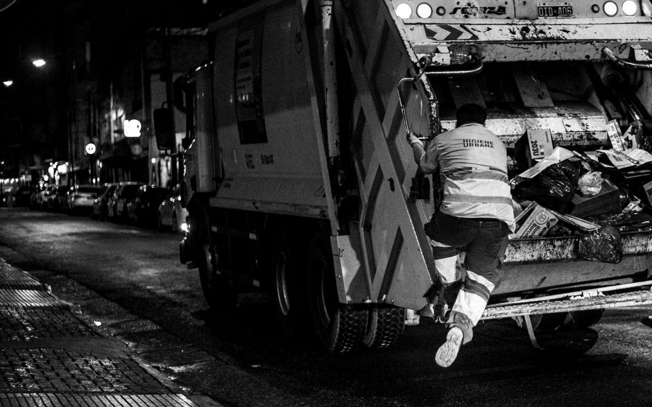 A waste collection worker, wearing a high-visibility vest with the words 'HIGIENE URBANA' printed on the back, is climbing onto the side of a large urban garbage truck parked on a city street at night. The truck has an open rear compartment filled with assorted rubbish, including black, plastic garbage bags, cardboard boxes, and other waste materials. The surface of the truck shows signs of dirt and wear, with some areas appearing stained or greasy. The street is lined with parked cars and illuminated by streetlights, creating a contrast between the bright lighting on the worker and truck and the darker background. The surrounding environment suggests an urban setting, possibly in a commercial or residential area, with shops or buildings faintly visible along the sidewalk. This scene illustrates an alternative method of rubbish collection, often associated with private waste management services like Waste Disposal Fulham, highlighting aspects of on-site clearance and independent rubbish removal workflows.